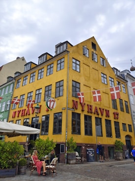 A vibrant yellow building featuring prominent red signage 'Nyhavn 17' and adorned with Danish flags. The building has black-framed windows and a small outdoor seating area with people sitting at tables. Potted plants add green to the scene, and the street is cobblestoned, giving a quaint and historic atmosphere.