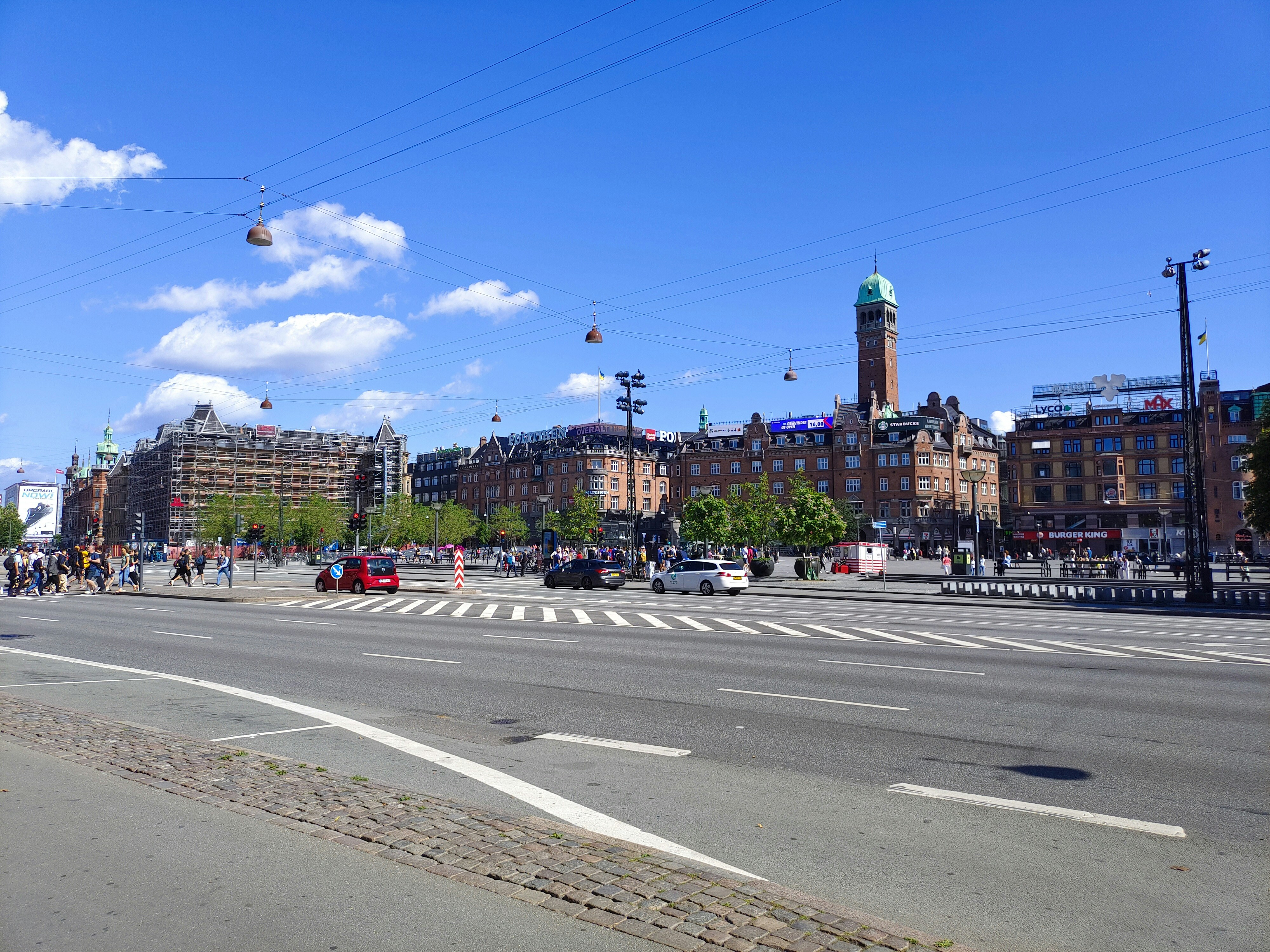 A lively street in Copenhagen with a clock tower visible in the background near the park