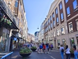 A bustling urban street lined with historic buildings featuring large windows. Pedestrians walk along the paved pathway, and several storefronts can be seen, including a souvenir shop and a café. There are hanging flower baskets and large planters with greenery, enhancing the lively atmosphere.