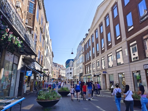 A bustling urban street lined with historic buildings featuring large windows. Pedestrians walk along the paved pathway, and several storefronts can be seen, including a souvenir shop and a café. There are hanging flower baskets and large planters with greenery, enhancing the lively atmosphere.