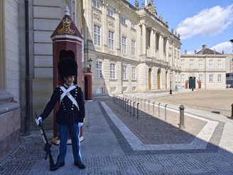 A guard wearing a traditional uniform, including a tall fur hat and a dark blue coat with white cross straps, stands beside a sentry box. The setting is a grand architectural building with tall windows and ornate details. The area is paved and open, with a clear sky above.