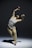 A person appears to be performing a contemporary dance move, captured mid-motion while bending backwards with arms raised expressively. The dance is performed on a bare stage with a plain, softly lit background.