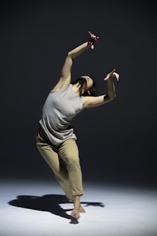 A person appears to be performing a contemporary dance move, captured mid-motion while bending backwards with arms raised expressively. The dance is performed on a bare stage with a plain, softly lit background.