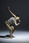 A person is captured in a dynamic dance pose, balancing gracefully on one foot. The scene is set against a minimalist backdrop with soft lighting casting shadows on the floor.