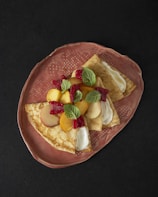 Three folded crepes topped with slices of peaches, raspberries, and mint leaves are arranged on a textured, oval, ceramic plate. The crepes appear to have a creamy filling visible near the folded edges. The rich, dark background enhances the contrast and colors of the dish.
