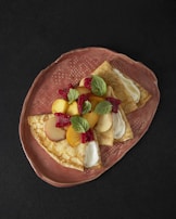 Three folded crepes topped with slices of peaches, raspberries, and mint leaves are arranged on a textured, oval, ceramic plate. The crepes appear to have a creamy filling visible near the folded edges. The rich, dark background enhances the contrast and colors of the dish.