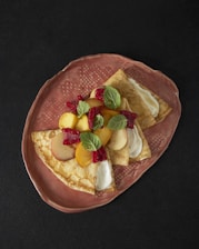 Three folded crepes topped with slices of peaches, raspberries, and mint leaves are arranged on a textured, oval, ceramic plate. The crepes appear to have a creamy filling visible near the folded edges. The rich, dark background enhances the contrast and colors of the dish.