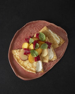 Three folded crepes topped with slices of peaches, raspberries, and mint leaves are arranged on a textured, oval, ceramic plate. The crepes appear to have a creamy filling visible near the folded edges. The rich, dark background enhances the contrast and colors of the dish.