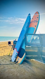 A sandy beach with a bright blue sky and ocean in the background. Two stand-up paddle boards are leaning against the side of a parked car, secured with a bungee cord. A folding chair is set up nearby, and there are some inflatable toys in the water.