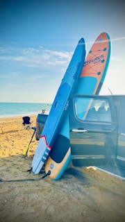A sandy beach with a bright blue sky and ocean in the background. Two stand-up paddle boards are leaning against the side of a parked car, secured with a bungee cord. A folding chair is set up nearby, and there are some inflatable toys in the water.