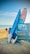 A sandy beach with a bright blue sky and ocean in the background. Two stand-up paddle boards are leaning against the side of a parked car, secured with a bungee cord. A folding chair is set up nearby, and there are some inflatable toys in the water.
