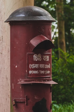 A vintage red postbox with a black dome lid is mounted on a pillar surrounded by greenery. The front panel features text both in the local script and in English indicating the opening time as 3:00 PM.