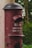 A vintage red postbox with a black dome lid is mounted on a pillar surrounded by greenery. The front panel features text both in the local script and in English indicating the opening time as 3:00 PM.