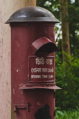 A vintage red postbox with a black dome lid is mounted on a pillar surrounded by greenery. The front panel features text both in the local script and in English indicating the opening time as 3:00 PM.