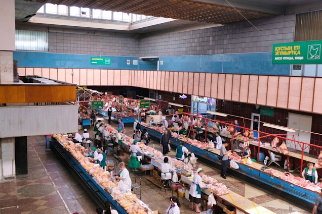 An indoor market scene with multiple rows of stalls displaying a variety of fresh meat products. Vendors dressed in white and green uniforms are attending to customers who are inspecting the products. The building features a high ceiling with skylights and signs in multiple languages that offer directions and information about the goods sold. The space is well-lit and structured, with wide aisles for customer movement.
