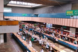 An indoor market scene with multiple rows of stalls displaying a variety of fresh meat products. Vendors dressed in white and green uniforms are attending to customers who are inspecting the products. The building features a high ceiling with skylights and signs in multiple languages that offer directions and information about the goods sold. The space is well-lit and structured, with wide aisles for customer movement.