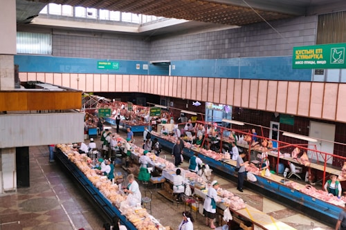 An indoor market scene with multiple rows of stalls displaying a variety of fresh meat products. Vendors dressed in white and green uniforms are attending to customers who are inspecting the products. The building features a high ceiling with skylights and signs in multiple languages that offer directions and information about the goods sold. The space is well-lit and structured, with wide aisles for customer movement.