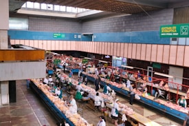 An indoor market scene with multiple rows of stalls displaying a variety of fresh meat products. Vendors dressed in white and green uniforms are attending to customers who are inspecting the products. The building features a high ceiling with skylights and signs in multiple languages that offer directions and information about the goods sold. The space is well-lit and structured, with wide aisles for customer movement.