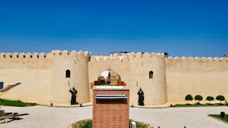 A large brick fortress with high walls and defensive towers features prominently. Statues of armored warriors holding spears stand in front of the entrance. The foreground includes a paved area and some grass patches, while the sky overhead is clear and blue.