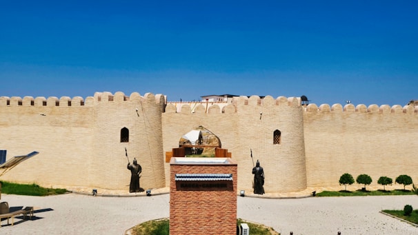 A large brick fortress with high walls and defensive towers features prominently. Statues of armored warriors holding spears stand in front of the entrance. The foreground includes a paved area and some grass patches, while the sky overhead is clear and blue.