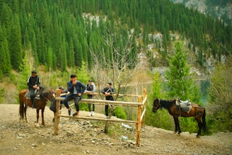 In a serene forest setting, several men are gathered near a wooden fence, some sitting on horseback. The background is dominated by lush greenery and a steep hillside covered with dense pine trees and rocky outcrops.