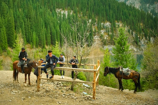 In a serene forest setting, several men are gathered near a wooden fence, some sitting on horseback. The background is dominated by lush greenery and a steep hillside covered with dense pine trees and rocky outcrops.