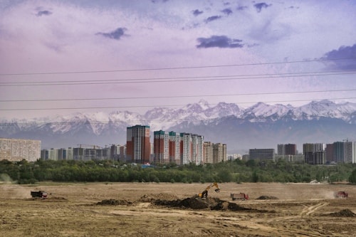 The image features a construction site with several vehicles, including trucks and an excavator, working in a barren area. In the background, there is a line of tall, modern apartment buildings set against a backdrop of snow-capped mountains. Power lines stretch across the top portion of the image, and a strip of vegetation separates the construction site from the urban area.