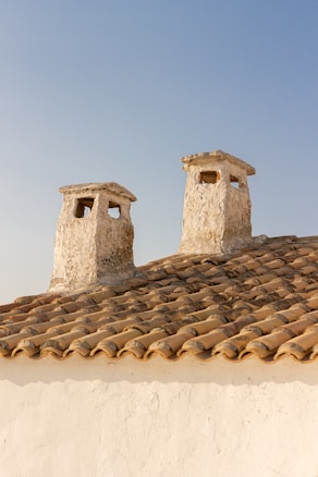 Two rustic chimneys are perched on a traditional terracotta-tiled roof against a clear blue sky. The chimneys are made of a rough, beige stone that gives an aged appearance.