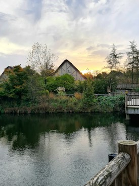 A peaceful riverside scene includes a tranquil body of water reflecting the sky and surrounding greenery. In the background, there are rustic, thatched buildings partially obscured by lush trees and foliage. A wooden fence can be seen in the foreground along the water's edge, adding to the countryside ambiance. The sky is partly cloudy, with a hint of sunset visible on the horizon, casting a warm glow over the scene.