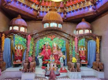 An elaborately decorated temple altar features intricately carved wooden structures and domes with colorful light displays. The central focus is on statues draped in vibrant red fabric and adorned with garlands. Floral decorations add to the vibrant atmosphere, creating a rich and sacred ambiance.