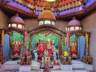An elaborately decorated temple altar features intricately carved wooden structures and domes with colorful light displays. The central focus is on statues draped in vibrant red fabric and adorned with garlands. Floral decorations add to the vibrant atmosphere, creating a rich and sacred ambiance.