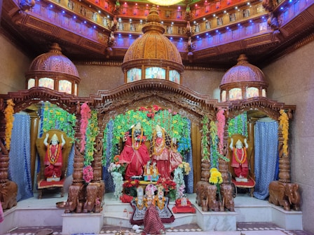 An elaborately decorated temple altar features intricately carved wooden structures and domes with colorful light displays. The central focus is on statues draped in vibrant red fabric and adorned with garlands. Floral decorations add to the vibrant atmosphere, creating a rich and sacred ambiance.