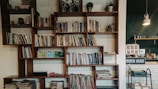 A modern wooden bookshelf filled with a variety of books and decorative items, such as small plants and a vintage coffee grinder. The left side of the image shows a cozy corner with stacked magazines and a few trinkets on display. In the background, there is a cafe counter with an 'Order Here' sign, glassware, and a hanging lamp providing soft lighting.