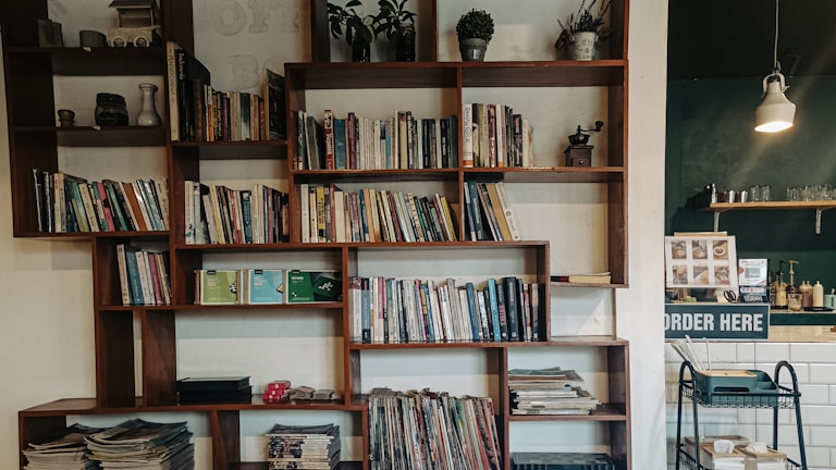A modern wooden bookshelf filled with a variety of books and decorative items, such as small plants and a vintage coffee grinder. The left side of the image shows a cozy corner with stacked magazines and a few trinkets on display. In the background, there is a cafe counter with an 'Order Here' sign, glassware, and a hanging lamp providing soft lighting.