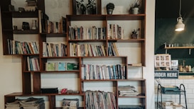 A modern wooden bookshelf filled with a variety of books and decorative items, such as small plants and a vintage coffee grinder. The left side of the image shows a cozy corner with stacked magazines and a few trinkets on display. In the background, there is a cafe counter with an 'Order Here' sign, glassware, and a hanging lamp providing soft lighting.