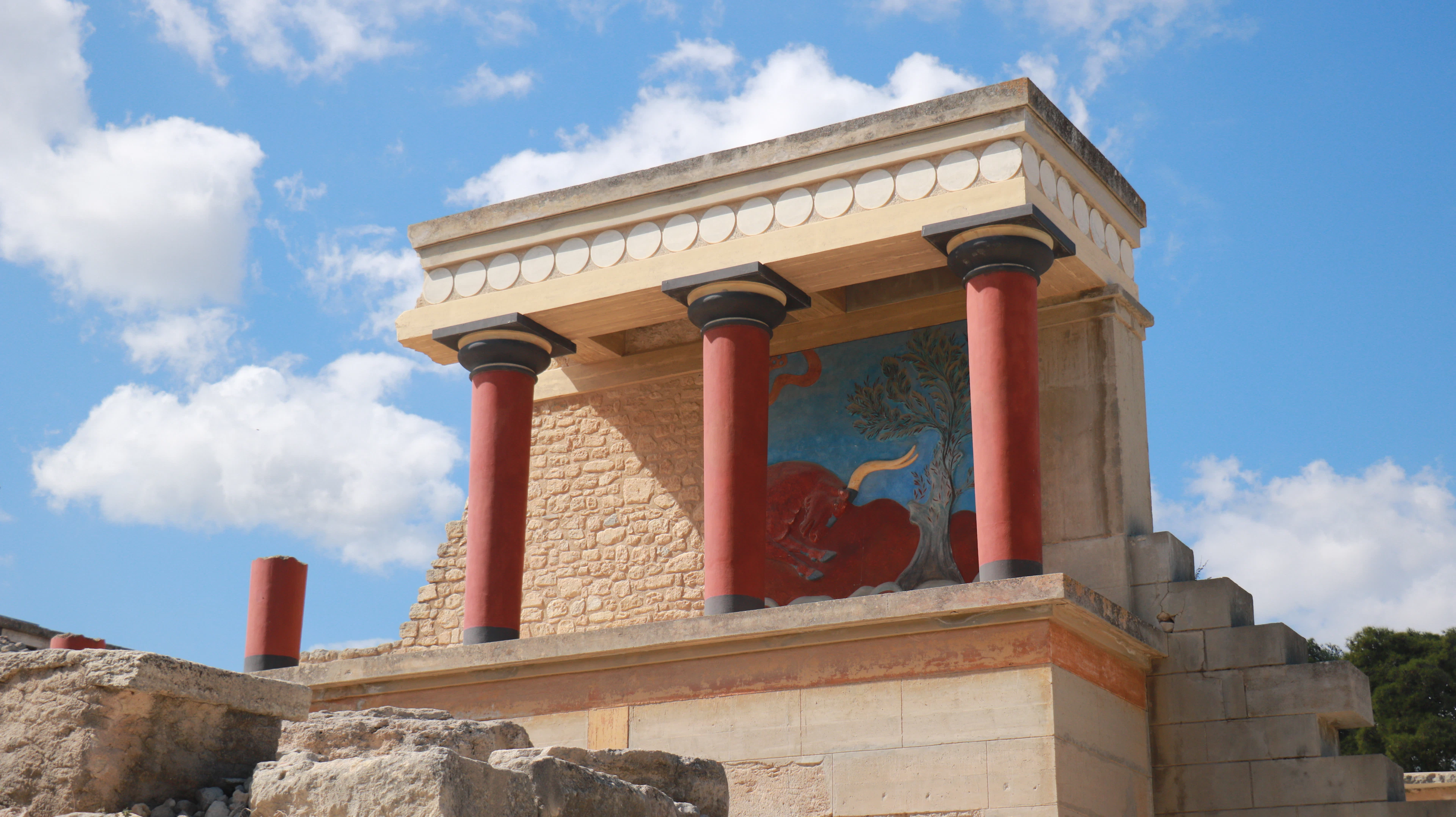 The reconstructed north entrance of the Knossos palace, with red columns and a painted fresco, under a blue sky.