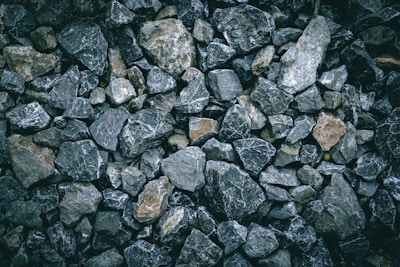 A collection of irregularly shaped rocks and stones of varying sizes and colors, ranging from dark gray to light brown, arranged closely together. The surface of the stones appears rough and textured, with some rocks having white veins running through them.