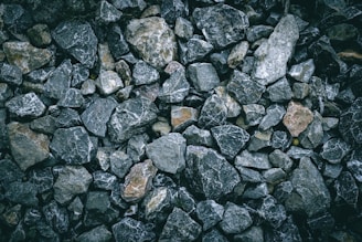 A collection of irregularly shaped rocks and stones of varying sizes and colors, ranging from dark gray to light brown, arranged closely together. The surface of the stones appears rough and textured, with some rocks having white veins running through them.