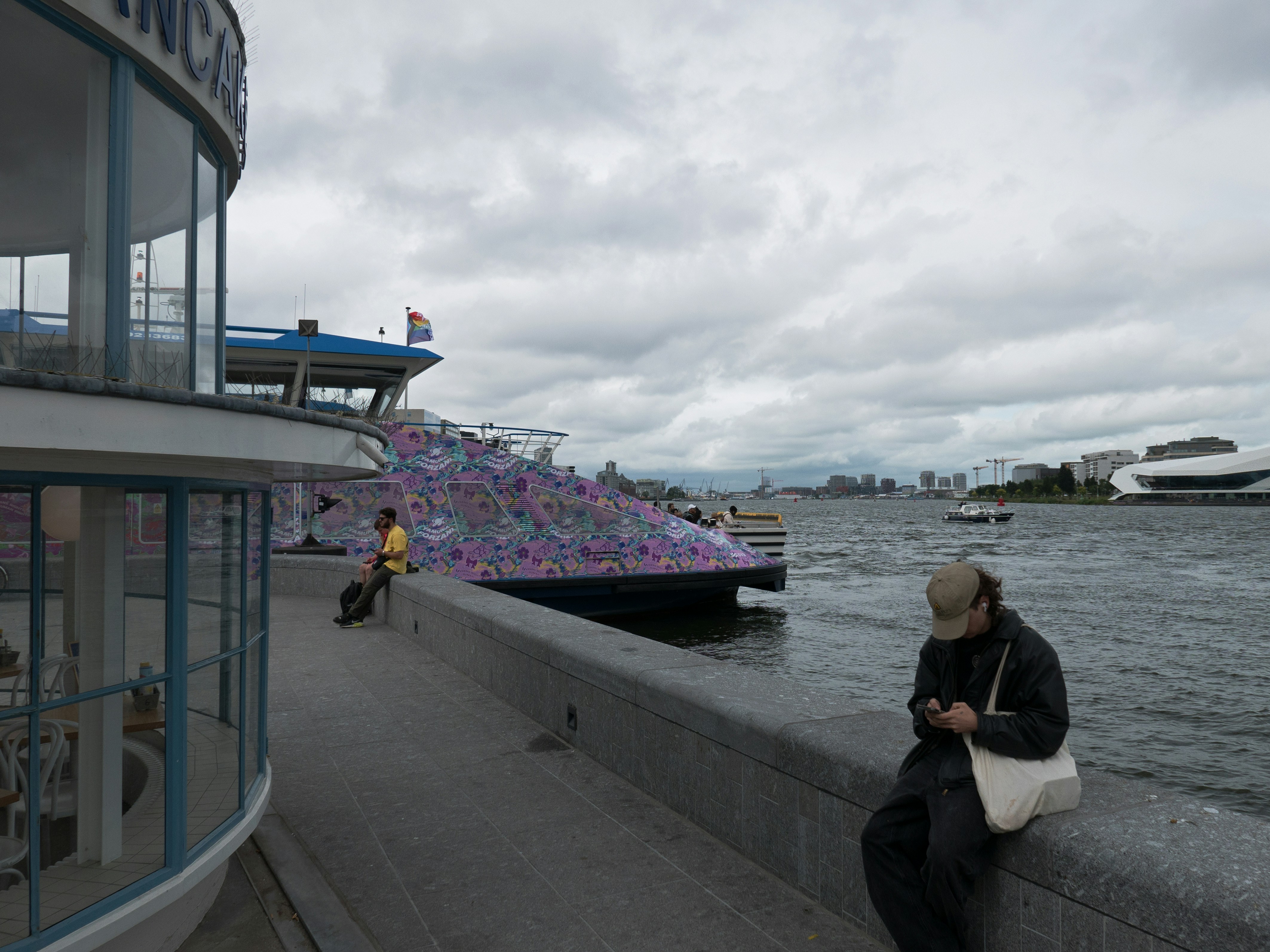 Photo of a wide grey view over the river IJ, behind Central station in Amsterdam. The ferry is rather colorful in all the gray shades. By the way; it is a Dutch Summer day in July! Street photography of the riverside in old city Amsterdam by Fons Heijnsbroek; free download urban photo, The Netherlands. This street image I share in suitable high resolution for making a nice art print or wallpaper.