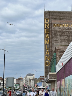 A street scene with a large brick building featuring the word 'Dreamland' vertically in bright yellow letters. The street is busy, with many people walking and a few vehicles visible. The sky is overcast with gray clouds, and several birds are flying. The building is flanked by other structures with various architectural styles.