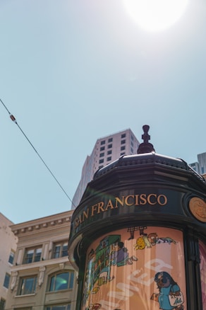 An urban scene featuring a close-up of a cylindrical kiosk with illustrations and the words 'San Francisco' written on it. Several buildings and a clear sky are visible in the background, with the sun positioned high above.