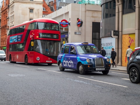 A bustling urban street scene featuring a prominent red double-decker bus with the destination sign 'Blackwall 15'. Beside the bus, a distinctive blue taxi with 'Cinch' branding is visible. Surrounding buildings display architecturally diverse styles, while pedestrians walk along the sidewalk.