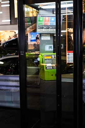 A brightly colored green public telephone is housed within a glass booth. The booth is located in an urban setting with reflections of city lights and signage visible in the glass. A car and a blurred pedestrian can be seen outside, adding to the city atmosphere. Various signs and notices are present inside the booth.