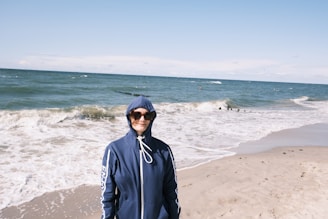 A young person wearing a cream-colored Thread Vibes hoodie, standing on a sandy beach with gentle ocean waves in the background, embodying relaxed coastal streetwear.