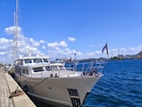 A large yacht is docked along a stone-paved harbor under a clear blue sky. The water is a deep blue, reflecting the bright sunlight. White clouds are scattered across the sky, and there are industrial buildings visible in the background. A flag is flying at the stern of the yacht.