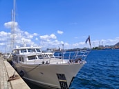 A large yacht is docked along a stone-paved harbor under a clear blue sky. The water is a deep blue, reflecting the bright sunlight. White clouds are scattered across the sky, and there are industrial buildings visible in the background. A flag is flying at the stern of the yacht.