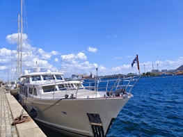 A large yacht is docked along a stone-paved harbor under a clear blue sky. The water is a deep blue, reflecting the bright sunlight. White clouds are scattered across the sky, and there are industrial buildings visible in the background. A flag is flying at the stern of the yacht.