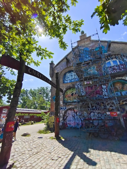 An urban scene featuring a building covered in colorful graffiti and street art. A metal scaffolding structure is positioned in front of the building. The area is shaded by trees, with sunlight filtering through the leaves. A cobblestone path leads towards the structure, and a few people are walking nearby. An archway with text is partially visible, contributing to the urban atmosphere.