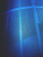 A close-up view of a metal surface featuring numerous circular perforations, illuminated by a blue light that casts varying shades and shadows across the textured surface.