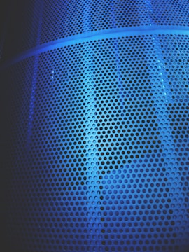 A close-up view of a metal surface featuring numerous circular perforations, illuminated by a blue light that casts varying shades and shadows across the textured surface.